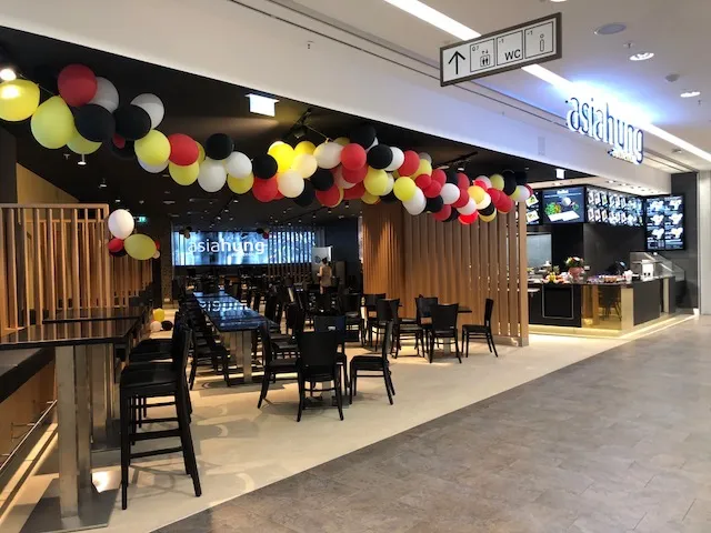 Innenansicht eines asiatischen Restaurants mit schwarzen Stühlen, langen Tischen und bunten Luftballons in Gelb, Rot, Weiß und Schwarz über der Sitzgelegenheit.