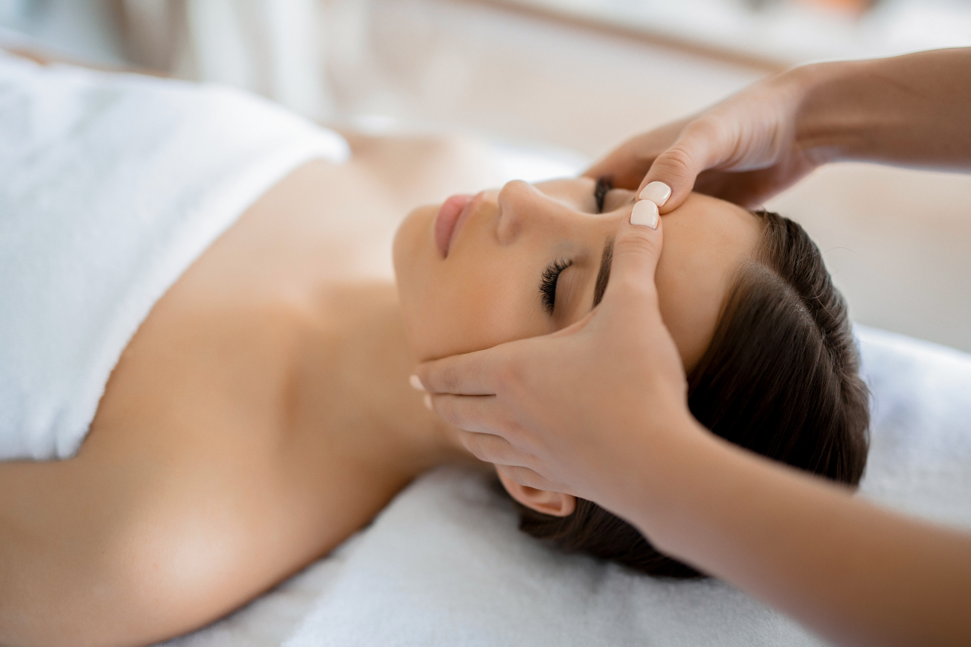 Woman receiving a facial massage while lying on a spa bed covered with a white towel.