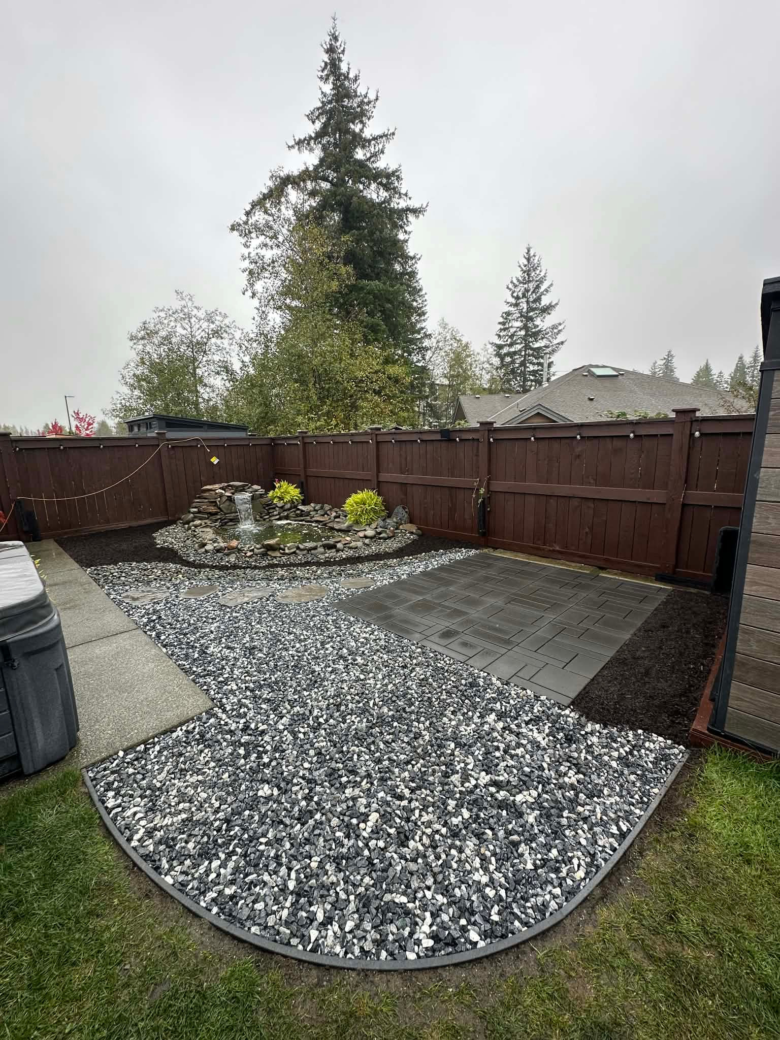 Backyard pond with a small waterfall, gravel landscaping and a paver pad beside a brown wood fence.