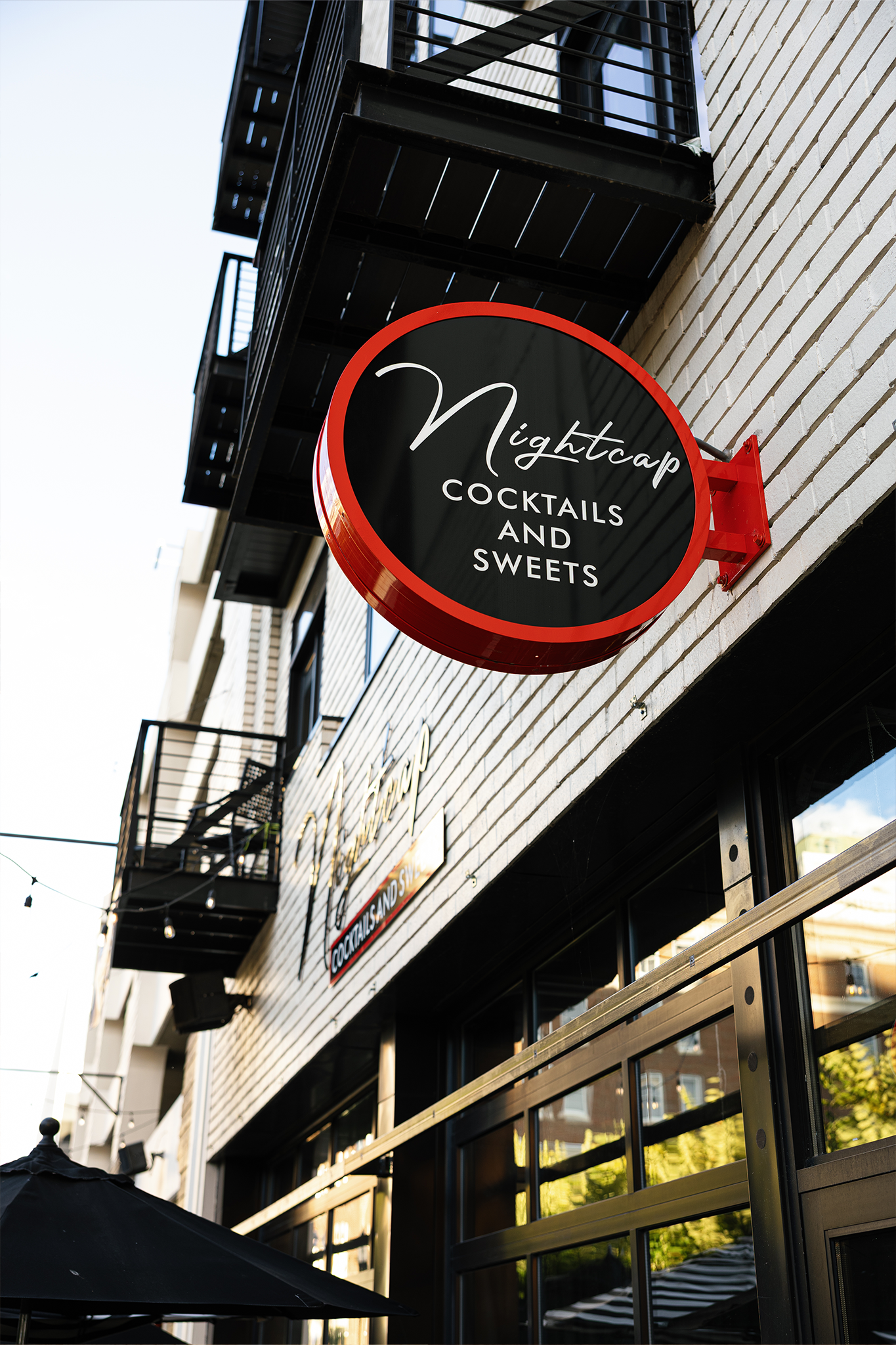 “Exterior sign for Nightcap Cocktails and Sweets mounted on a white brick building with black metal balconies above.”
