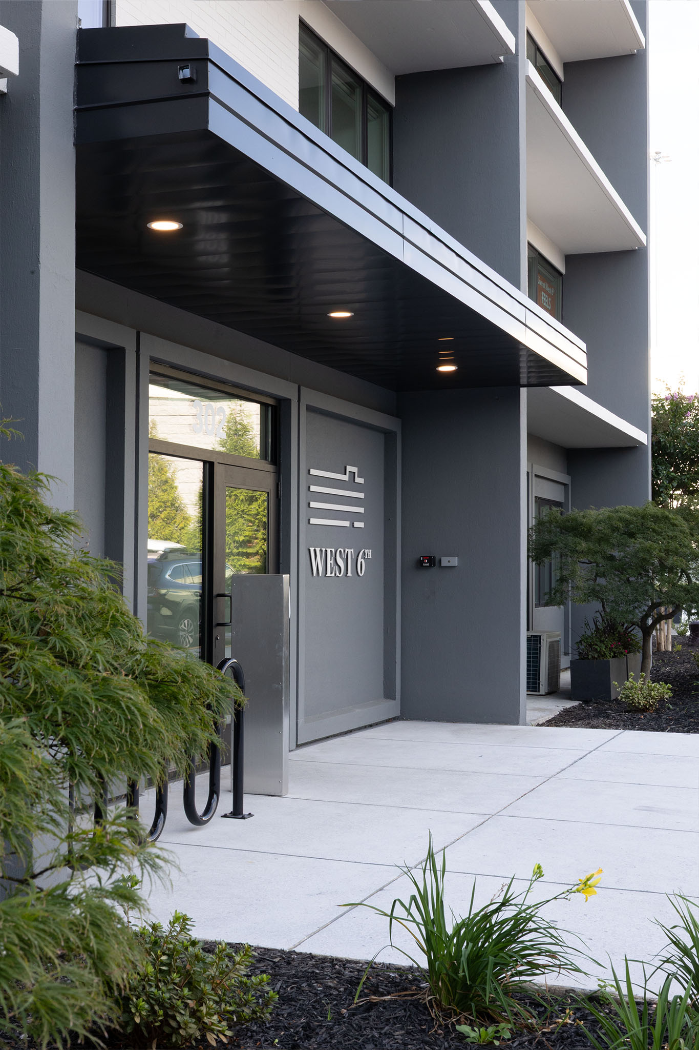 “Exterior entrance of West 6th building with modern gray façade, glass door, and sign reading ‘West 6th,’ surrounded by greenery.”