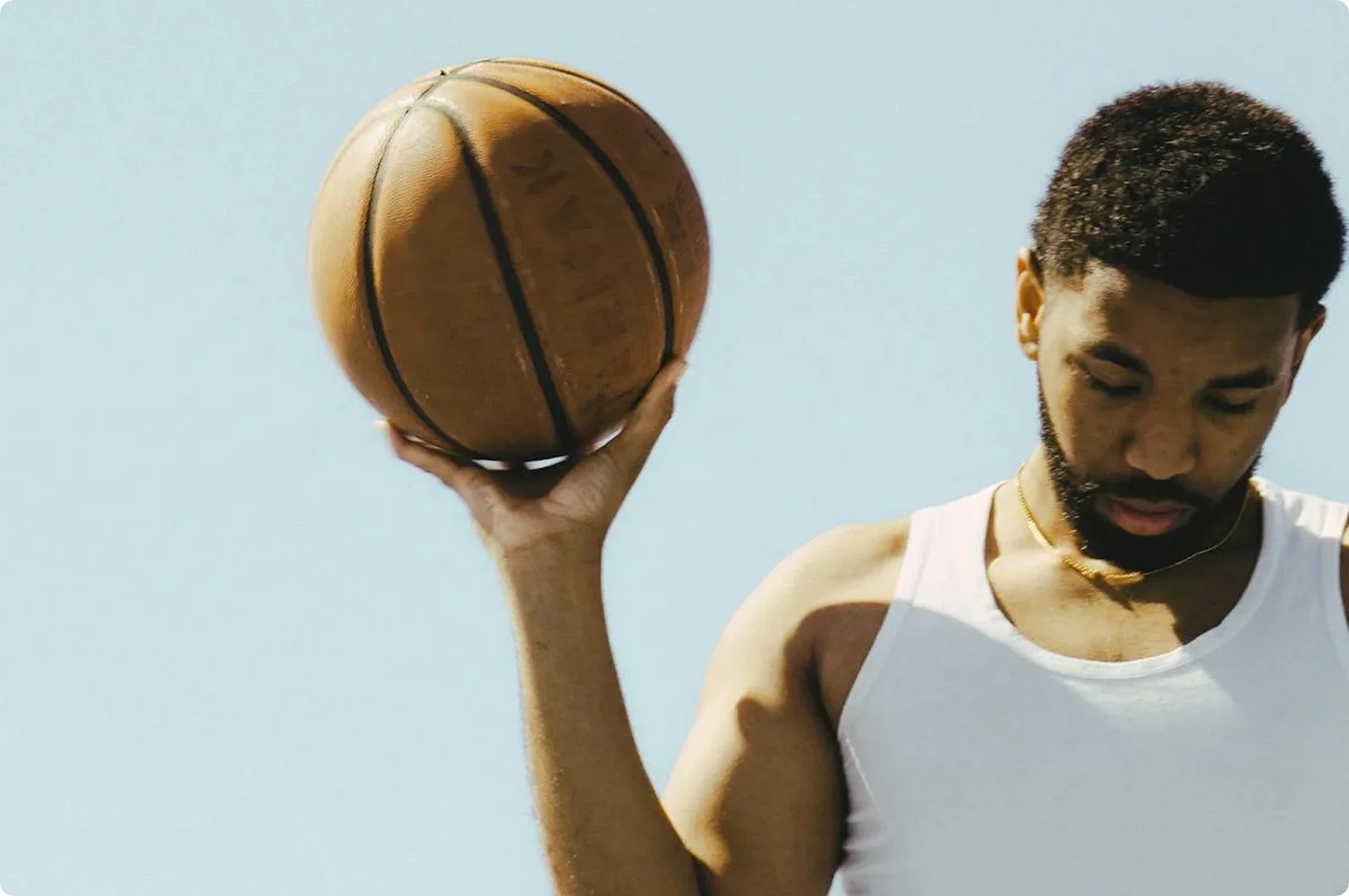 Man in a white tank top holding a basketball with one hand against a clear blue sky.
