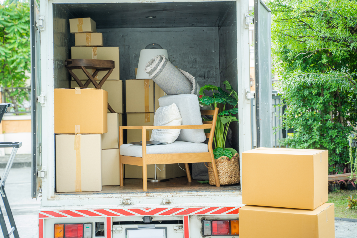 Open moving truck loaded with stacked cardboard boxes, a chair with cushions, a rolled rug, a lamp, and a potted plant.