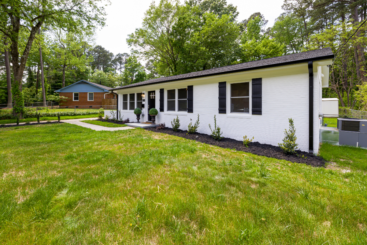 Single-story white brick house with black shutters, a small front porch with potted plants, and a grassy yard surrounded by trees.