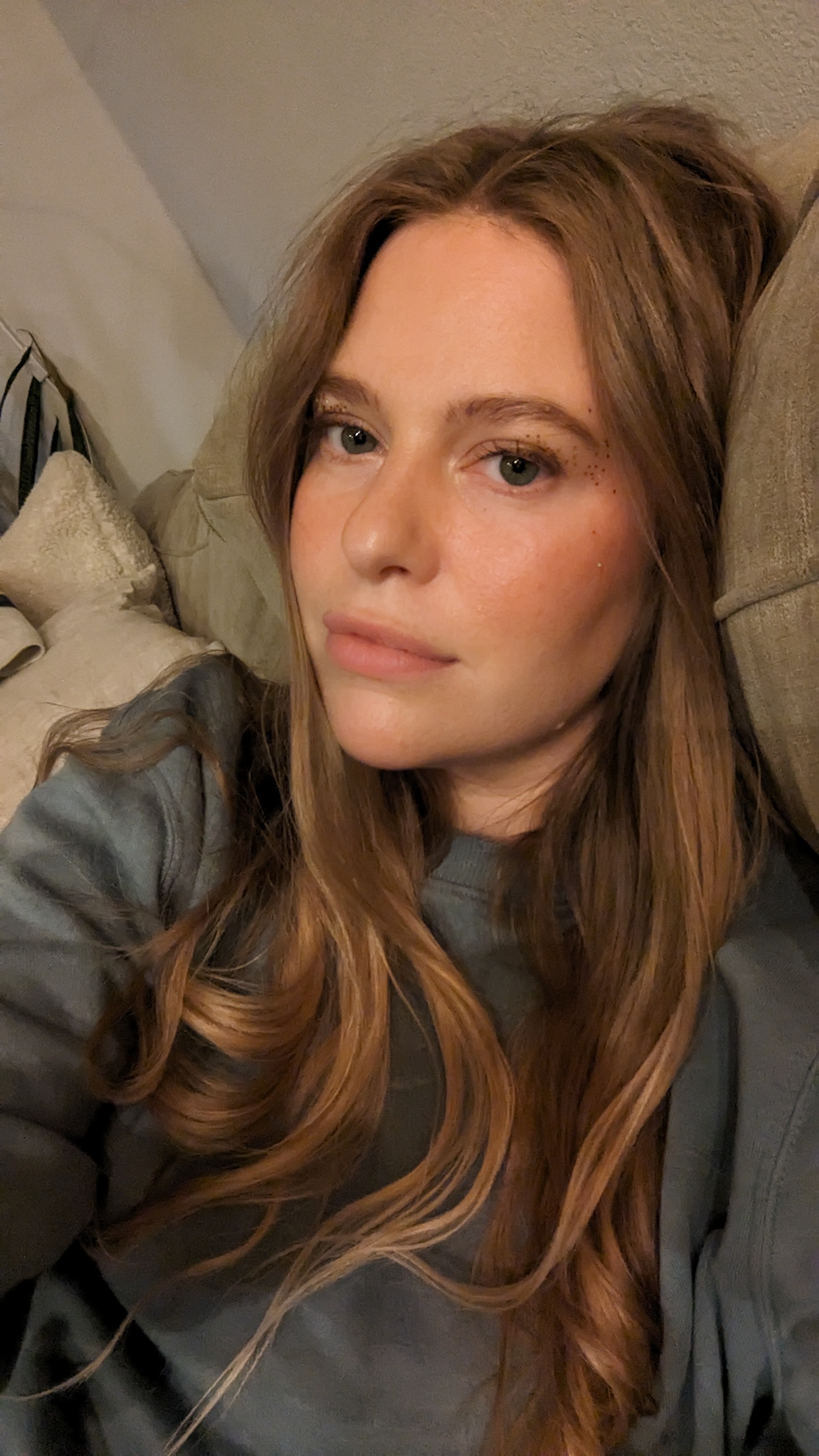 Close-up of a young woman with long light brown hair reclining on a couch with a neutral expression.