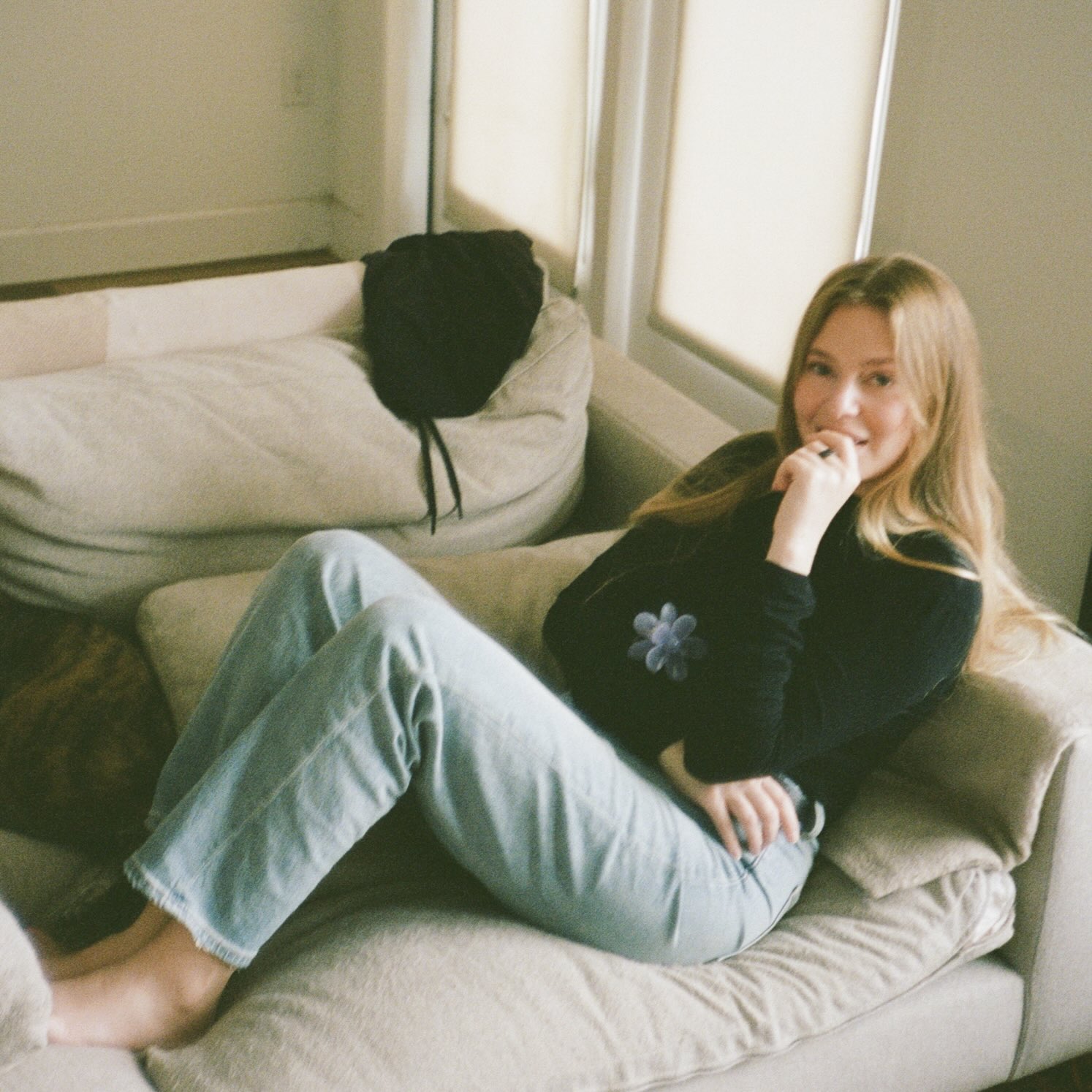 Smiling woman with long blonde hair sitting barefoot on a beige couch wearing light blue jeans and a black top.