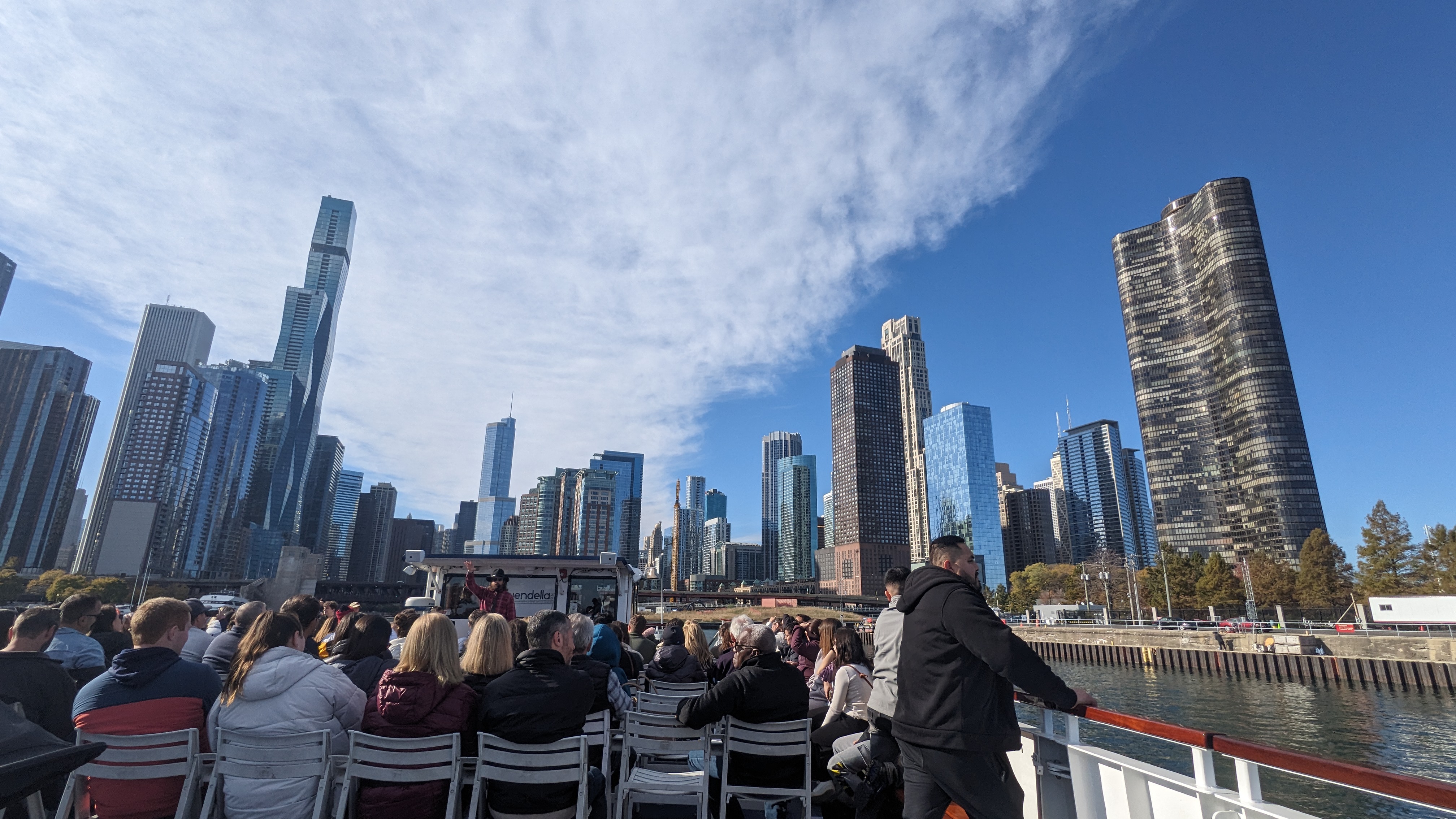 Group of people seated on a boat with a city skyline of tall modern buildings and a partly cloudy blue sky in the background.