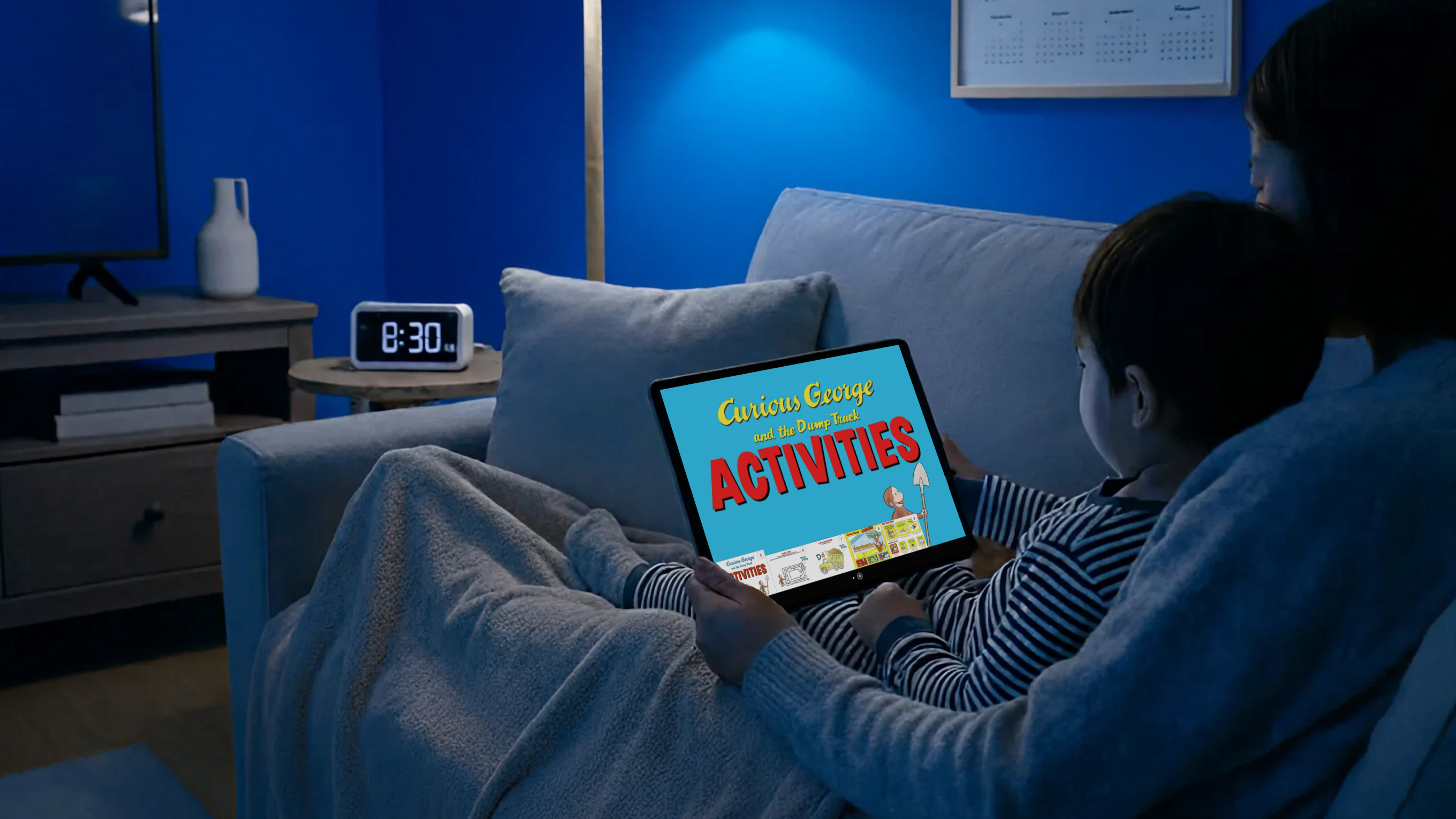 A child and an adult sitting on a couch at night looking at a tablet displaying 'Curious George and the Dump Truck ACTIVITIES'.