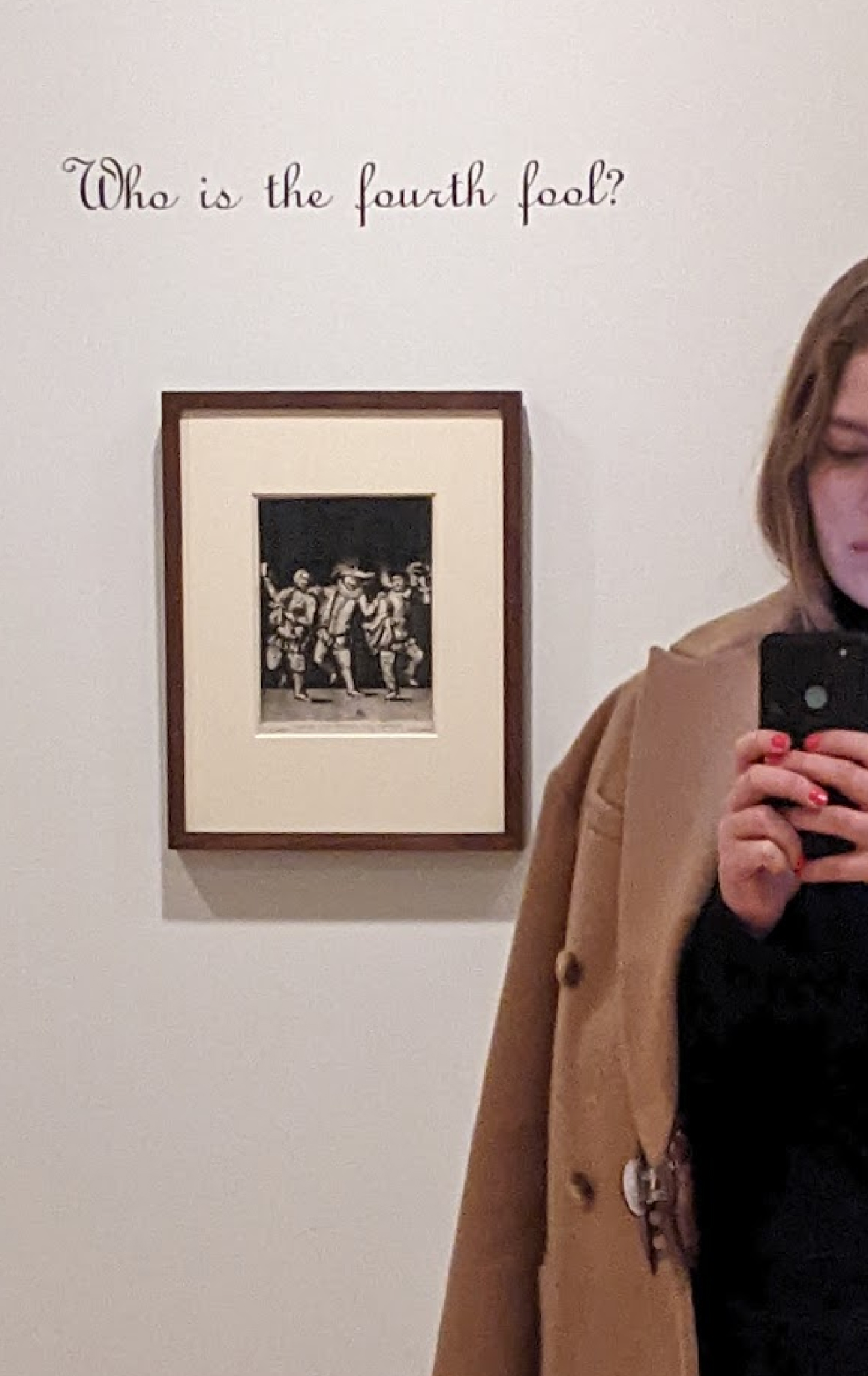 Person taking a selfie in a mirror showing a wall with a framed black and white artwork of dancing figures and text above reading 'Who is the fourth fool?'