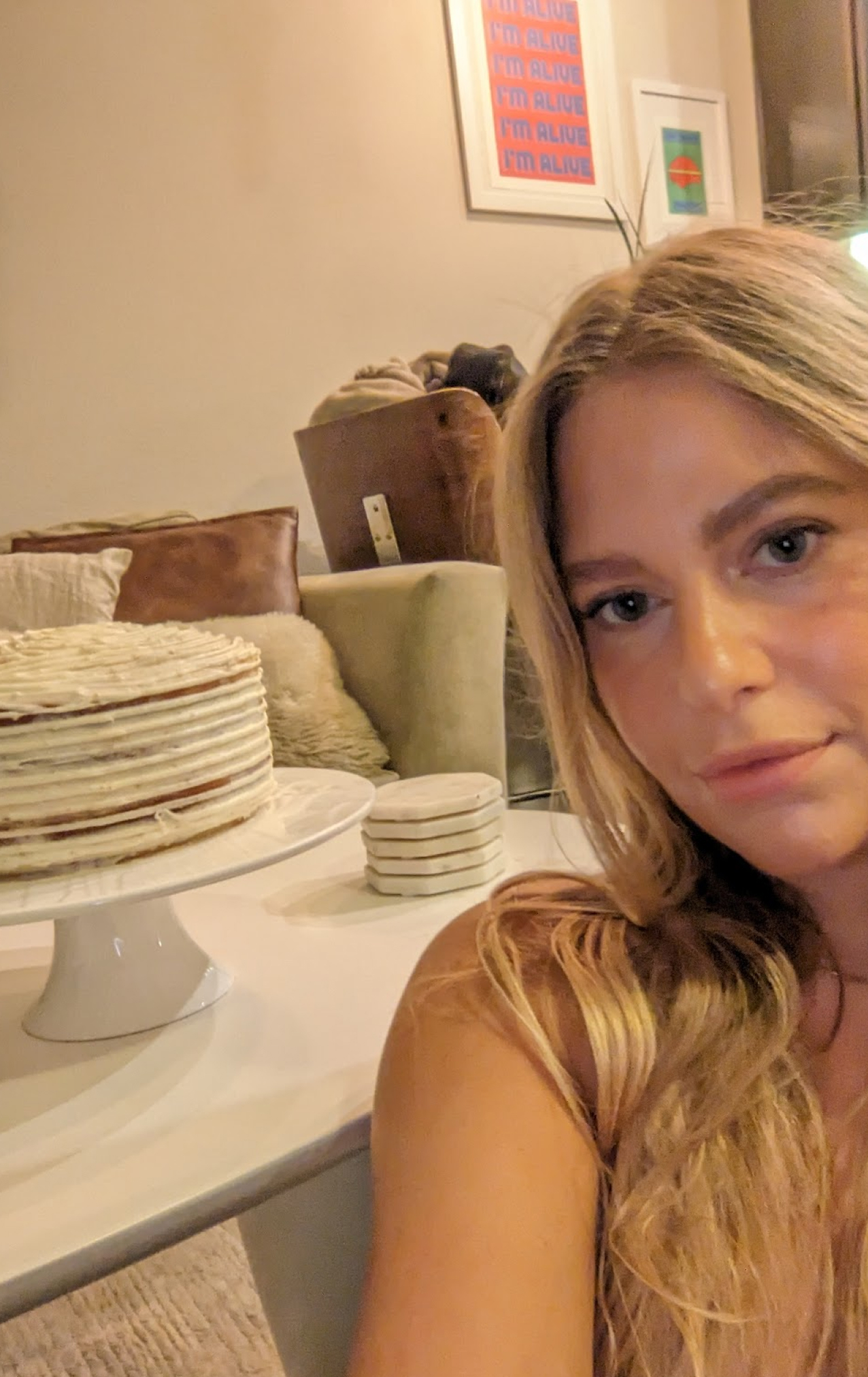 Close-up of a woman with long blonde hair taking a selfie indoors next to a white cake on a cake stand and stacked coasters on a table.
