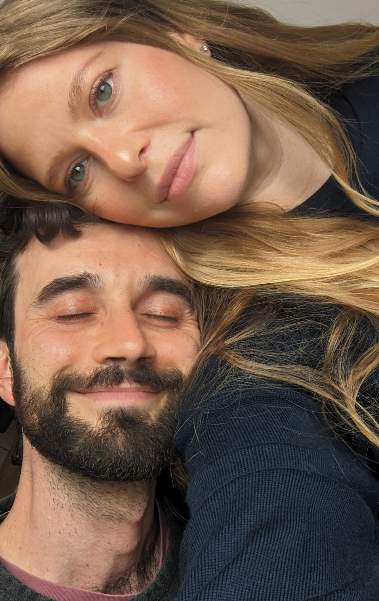 Close-up of a smiling man with closed eyes and a woman with green eyes leaning her head on his, both with light skin and the woman having long blonde hair.