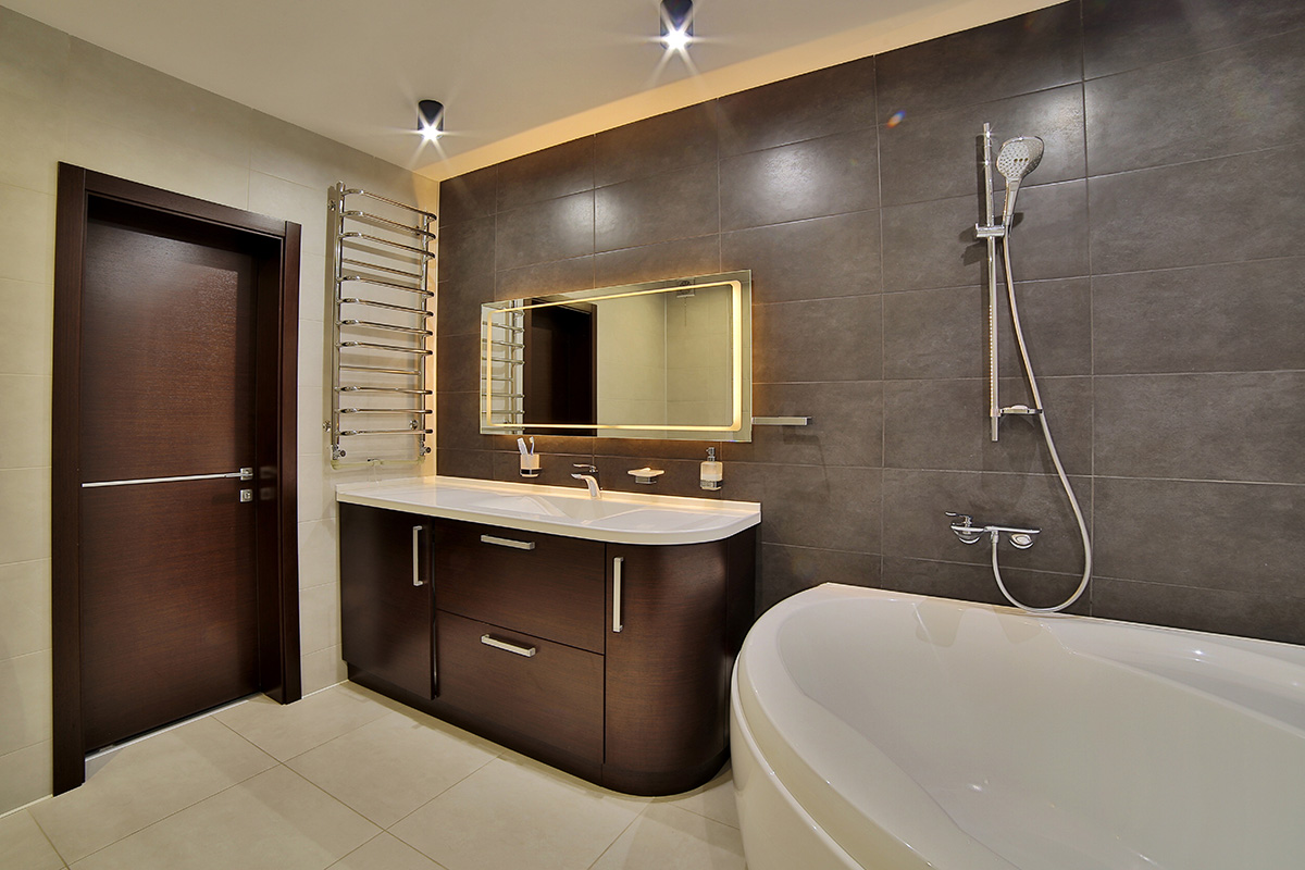 Modern bathroom with dark wood vanity, illuminated rectangular mirror, white corner bathtub, and gray tiled wall with showerhead.