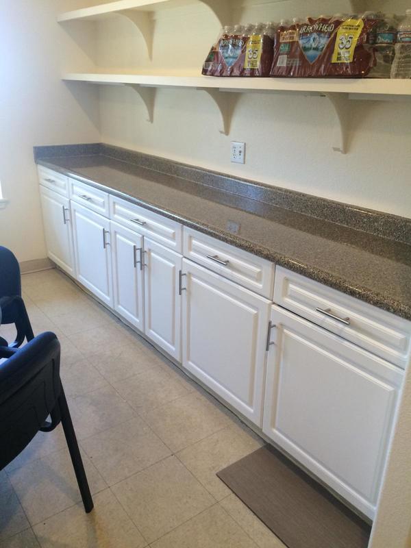 Kitchen area with white cabinets under a granite countertop and two shelves above holding packs of bottled water.