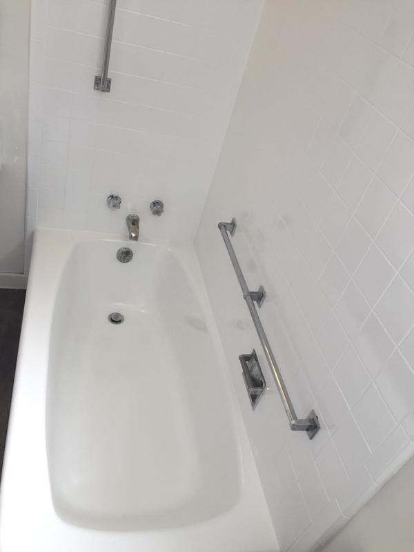 Clean white bathtub with chrome faucet, two handles, and a metal grab bar mounted on white tiled wall.