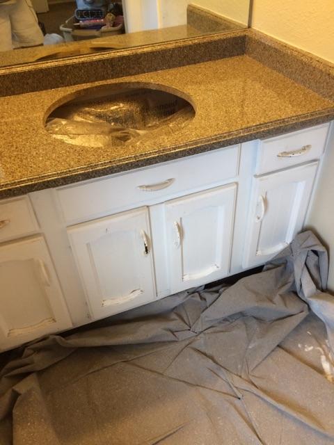 White bathroom cabinet with chipped paint and a granite countertop with an open sink hole, covered in plastic, with a gray protective cloth draped on the floor.