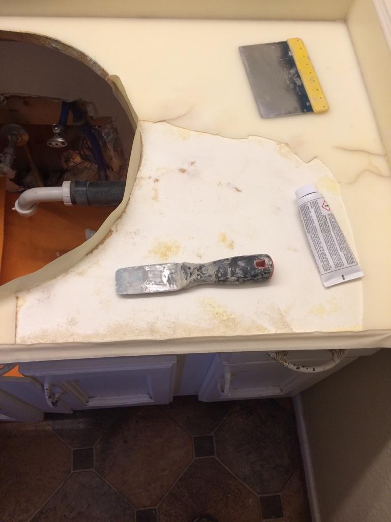Countertop with a cutout hole exposing plumbing and tools including a putty knife, a scraper, and a small tube of adhesive or filler.