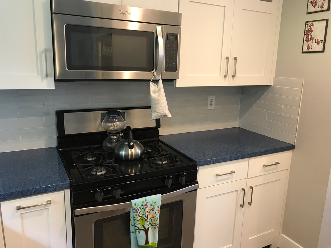 Modern kitchen stove with a stainless steel microwave above, a kettle and coffee maker on the stovetop, and a decorative towel hanging on the oven door.