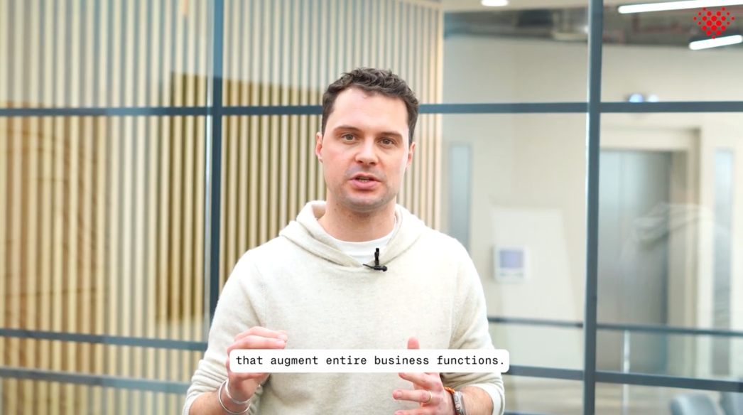 Man in white hoodie speaking indoors with subtitle text reading 'that augment entire business functions.'
