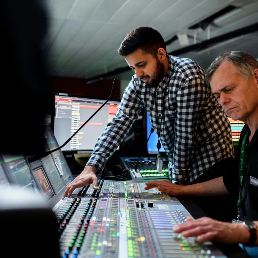 Two men adjusting controls on a large audio mixing console in a studio with screens in the background.