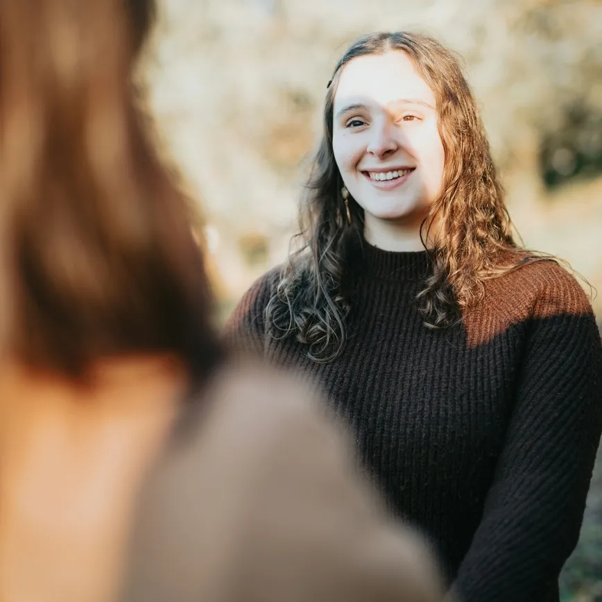 Smiling woman with curly hair wearing a dark sweater talking to another person outdoors.