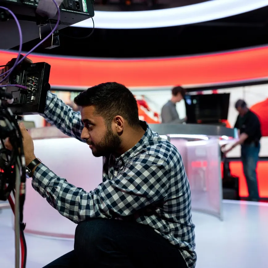 Man in checkered shirt adjusting camera equipment in a modern media studio with blurred staff in background.