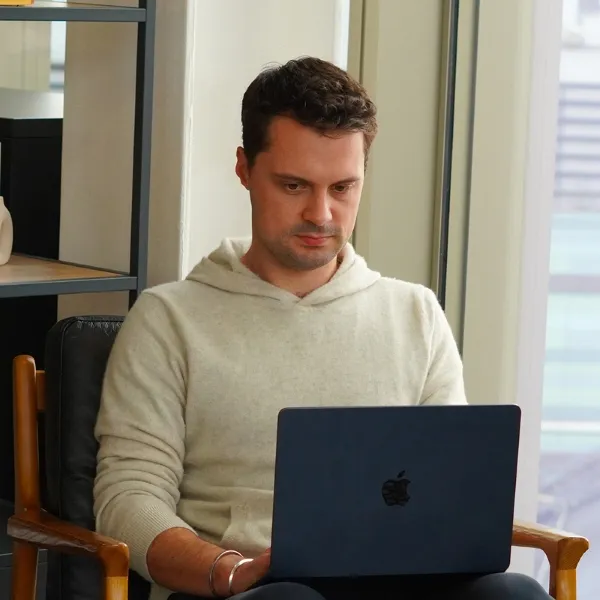 Two men sitting at a table with laptops, smiling and talking near large windows and green plants.