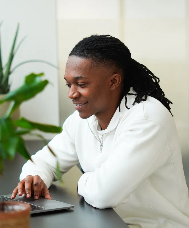 Smiling man with dreadlocks wearing white sweater using laptop at a desk with plants nearby.