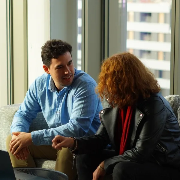 Two men sitting at a table with laptops, smiling and talking near large windows and green plants.