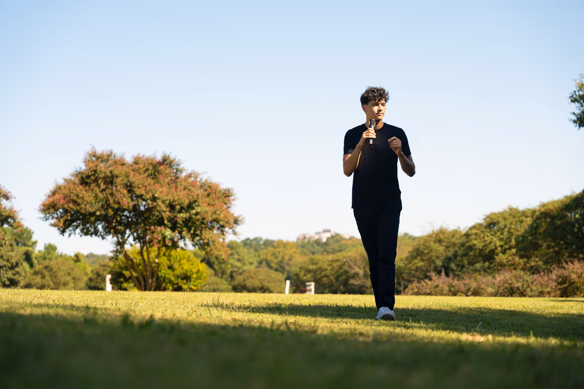 Young man in black clothing holding a microphone walking on grass in a park with trees in the background under a clear sky.