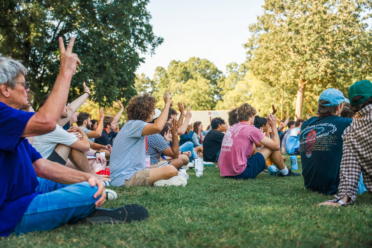 Group of people sitting on grass in a park, raising their hands during an outdoor event.