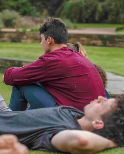 Young people relaxing outdoors on grass, one sitting with arms around knees and another lying down with hands behind head.