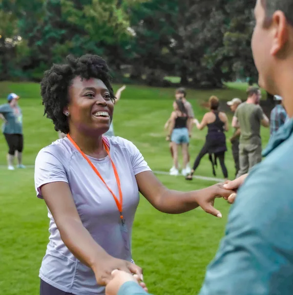 Smiling woman holding hands with a man outdoors on green grass with people in the background.