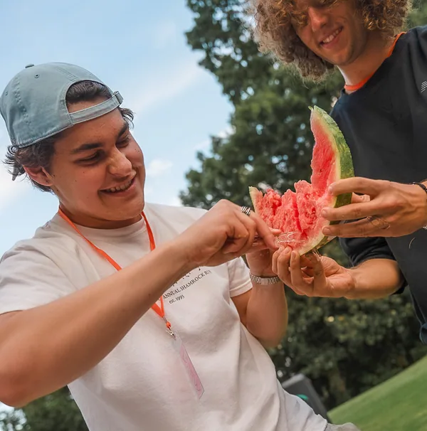Two young men outdoors sharing and eating a large slice of watermelon on a sunny day.