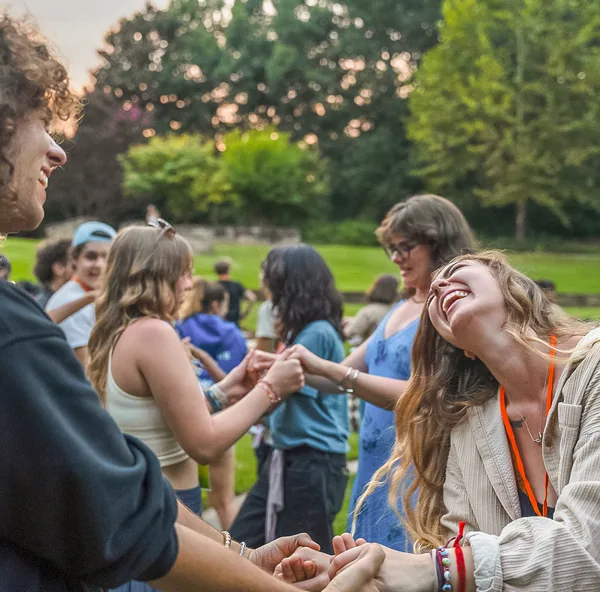 Young people laughing and holding hands in a circle outdoors during a social gathering.