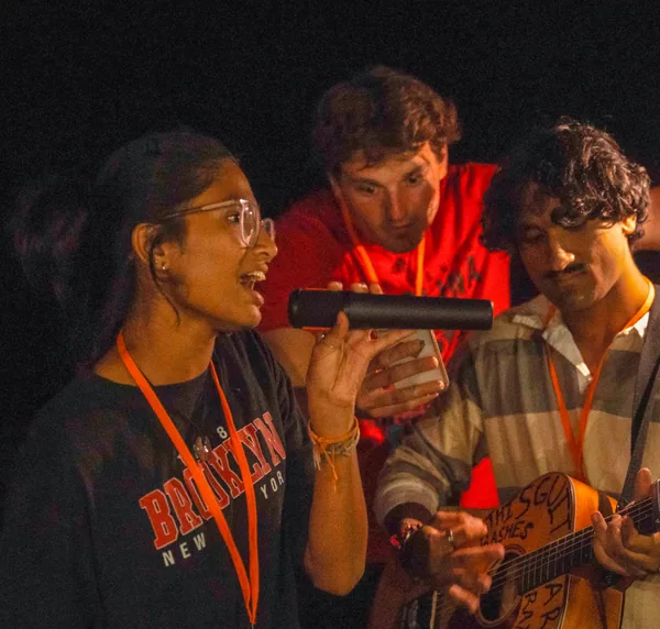 Young woman singing into a microphone while a man plays an acoustic guitar and another man looks at a phone in the background.