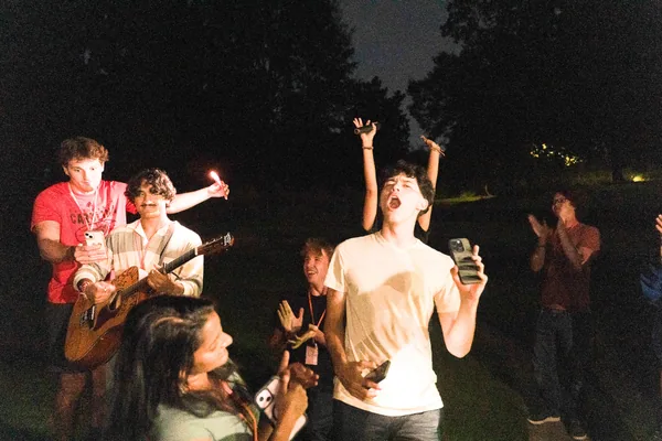 Group of young people enjoying a nighttime outdoor gathering with singing, guitar playing, and clapping.