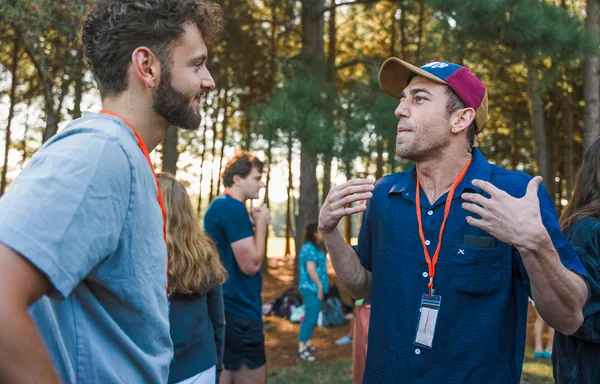 Two men with lanyards talking animatedly outdoors with other people and trees in the background.