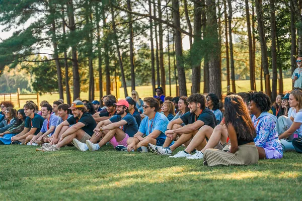 Group of young people sitting on grass outdoors, listening attentively with trees in the background.