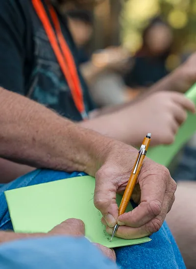 Person holding a pencil and writing on green paper while wearing a dark shirt with an orange lanyard.