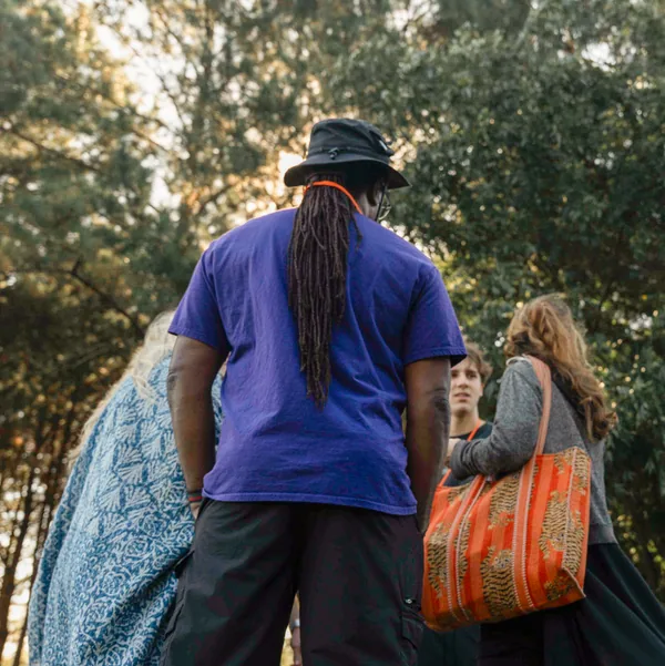Group of four people outdoors, with one person in a purple shirt and black hat facing away, and others engaged in conversation near trees.
