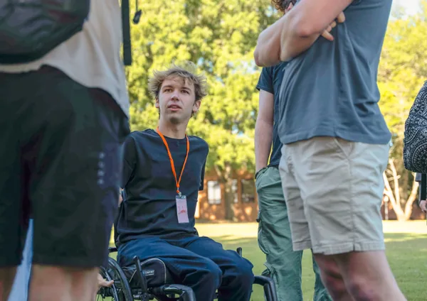 Young man in a wheelchair wearing a black sweater and orange lanyard talking with standing people outdoors on a sunny day.