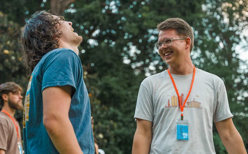 Two young men outdoors laughing and talking, one wearing a white t-shirt with glasses and a name tag on an orange lanyard, the other in a blue shirt.