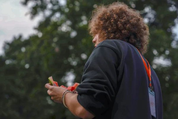 Person with curly hair wearing a dark sweatshirt and an orange lanyard, holding watermelon slices outdoors.
