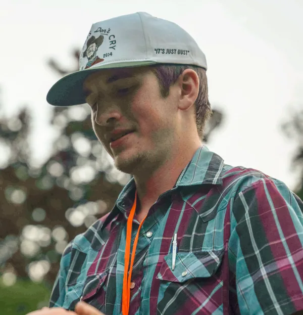 Man wearing a gray cap with text and a tattooed face logo, a plaid shirt in teal and burgundy, and an orange lanyard outdoors.
