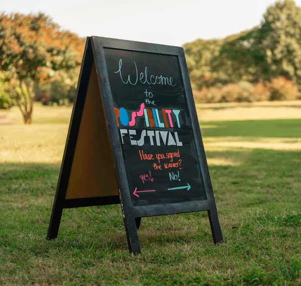 Chalkboard sign on grass welcoming visitors to the Possibility Festival, asking if they have signed the waiver with arrows pointing left for yes and right for no.