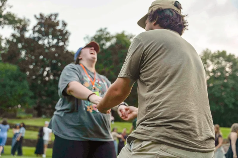 Two people outdoors holding hands and laughing with a blurred crowd and trees in the background.