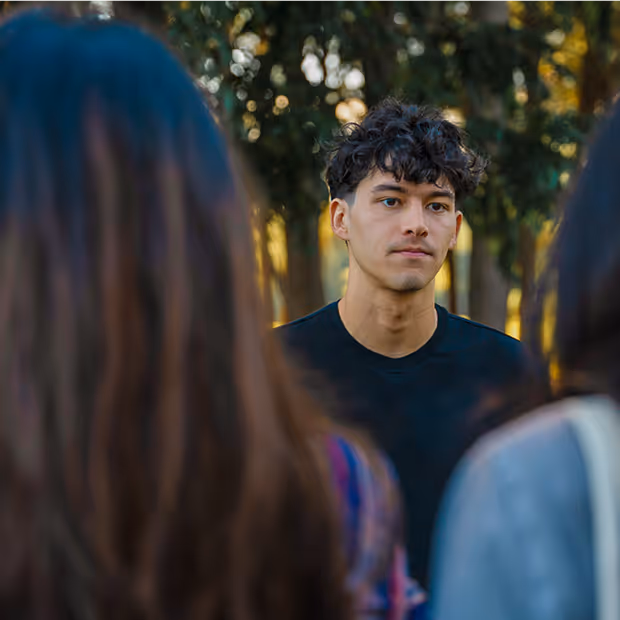 Young man with curly hair wearing a black shirt looking thoughtfully ahead, framed by blurred women in the foreground outdoors.