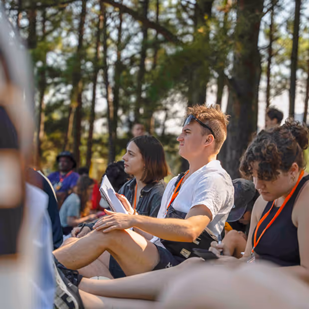 Young people sitting outdoors in a wooded area, attentively watching or listening, with one person holding a notebook.