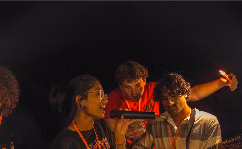 Young woman singing into a microphone with two men beside her, one holding a guitar and the other shining light from a phone.