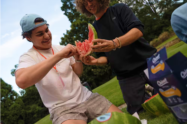 Two young people outdoors breaking apart a large slice of watermelon while sitting on grass in a park.